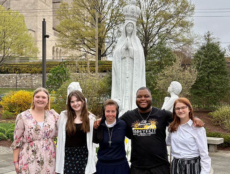 Three young woemn, one young man and a nun standing in front of religious statue