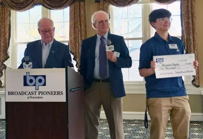 Young man holding up large check, next to two men in suits behind a podium with sign "Broadcast Pioneers of Philadelphia"