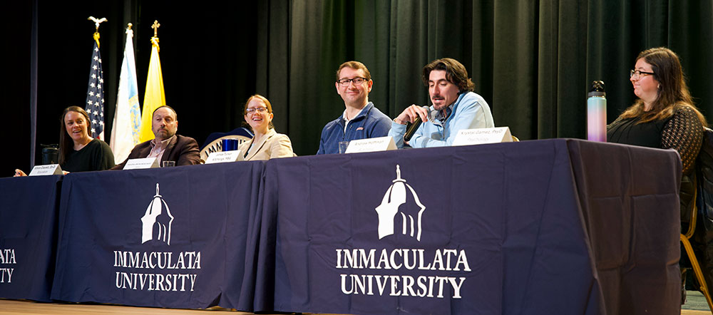 Six people sitting behind a table at a panel discussion. The table has a banner with image of dome and text Immaculata University